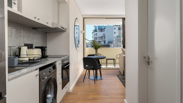 Cocina compacta con lavadora, zona de comedor con mesa y sillas, y vista panorámica de la ciudad desde un balcón.