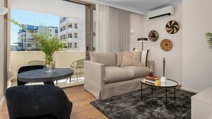 Una sala de estar moderna con un sofá, una mesa de centro con libros, una planta en maceta y vistas de un edificio a través de amplios ventanales.