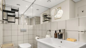 Un baño moderno con lavamanos, inodoro y espejo blancos. Hay repisas con artículos de aseo personal y un cesto de basura.