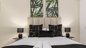 Un lit bien fait, avec des serviettes blanches pliées en rouleau, deux tables de chevet avec des lampes et une tête de lit ornée d’un motif de feuilles.