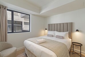 Een netjes opgemaakt bed met hoofdeinde, een nachtkastje met lamp, een stoel en een raam met gordijnen.