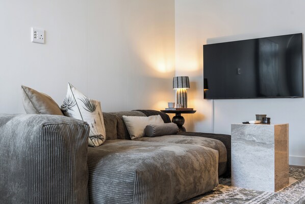 Een moderne woonkamer met een grijze bank, een bijzettafeltje, een flatscreen televisie en een kamerkleed met een patroon.