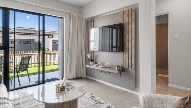 A modern living room with a large sliding glass door leading to a patio, a flat-screen TV mounted on the wall, and a small side table with a remote control. - Bliss Ballito | Comfort and Style 2x Bedroom (Dolphin Coast)