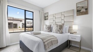 A modern bedroom with a large bed, a bedside table with a lamp, and a view of a building through a window. - Bliss Ballito | Comfort and Style 2x Bedroom (Dolphin Coast)