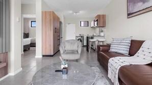 A modern living room with a brown leather sofa, a grey armchair, and a coffee table. A kitchen area is visible in the background with wooden cabinets and a dining table. - Bliss Ballito | Modern Apartment and Clubhouse (Dolphin Coast)