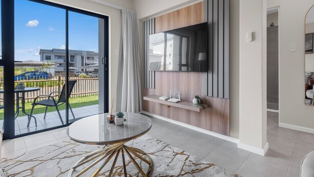 A modern living room with a large TV, a round table, and a sliding glass door leading to a balcony with outdoor furniture. - Bliss Ballito | Modern Apartment and Clubhouse (Dolphin Coast)