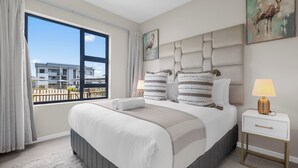 A modern bedroom with a large bed, a view of the outside balcony, and a nightstand with a lamp. - Bliss Ballito | Modern Apartment and Clubhouse (Dolphin Coast)