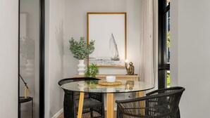 Een eetruimte met een glazen tafel, zwarte rieten stoelen, een kunstwerk van een zeilboot in een lijst, een potplant en een klein beeldje.