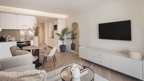 Un soggiorno moderno con divano, tavolino e televisore. Presenta un angolo cottura con pensili e un tavolo da pranzo.