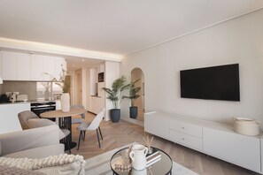 A modern living room with a sofa, coffee table, and TV. There is a kitchen area with cabinets and a dining table. - Sea Point Gem | Balcony and Central Location (Cape Town)