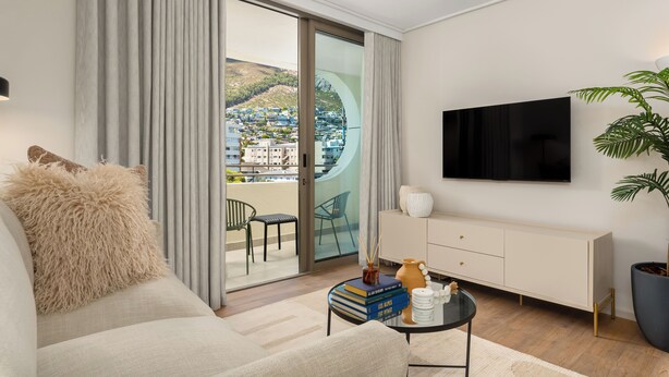 A modern living room with a sofa, a coffee table with books, a TV, and a view of a cityscape through a sliding glass door. - Dolce Vita | Designer Apartment | Lion's Head View (Cape Town)