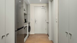 A narrow hallway with a kitchen area on the left, featuring a refrigerator, oven, and washing machine. A white door is at the end of the hallway. - Dolce Vita | Designer Apartment | Lion's Head View (Cape Town)