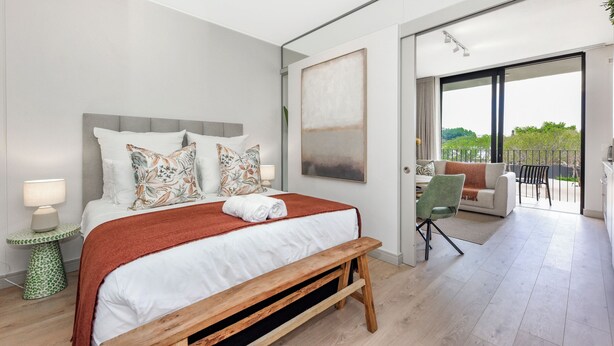 A modern bedroom with a large bed, a bench, a desk, and a view of the outdoors. - One Rosebank Premium Luxe Apartment (Johannesburg)