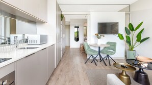 A modern kitchen with white cabinets, a built-in microwave, and a sink. There is a dining area with a round table and chairs, and a large potted plant. - One Rosebank Premium Luxe Apartment (Johannesburg)