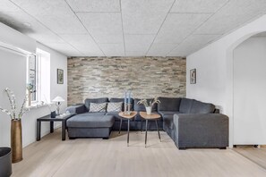 Ein modernes Wohnzimmer mit einem grauen Ecksofa, zwei kleinen Beistelltischen, einer Wand mit Dekoration und einem Fenster mit Ausblick.