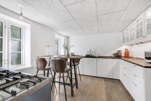 Eine moderne Küche mit weißen Schränken, einem Gasherd, einem Esstisch mit Stühlen und einem Fenster mit Tageslicht.