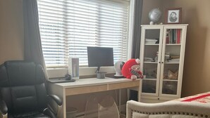A room with a desk, chair, and a bed. There is a window with blinds, a bookshelf, and a rug on the floor.