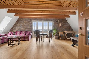 A loft-style room with a stone wall, wooden ceiling beams, a stone fireplace, a round table with chairs, a sofa, and a large window.