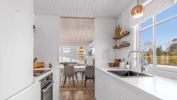 A modern kitchen with white cabinetry, a dining area with a table and chairs, a large window with a view, and a hanging light fixture.