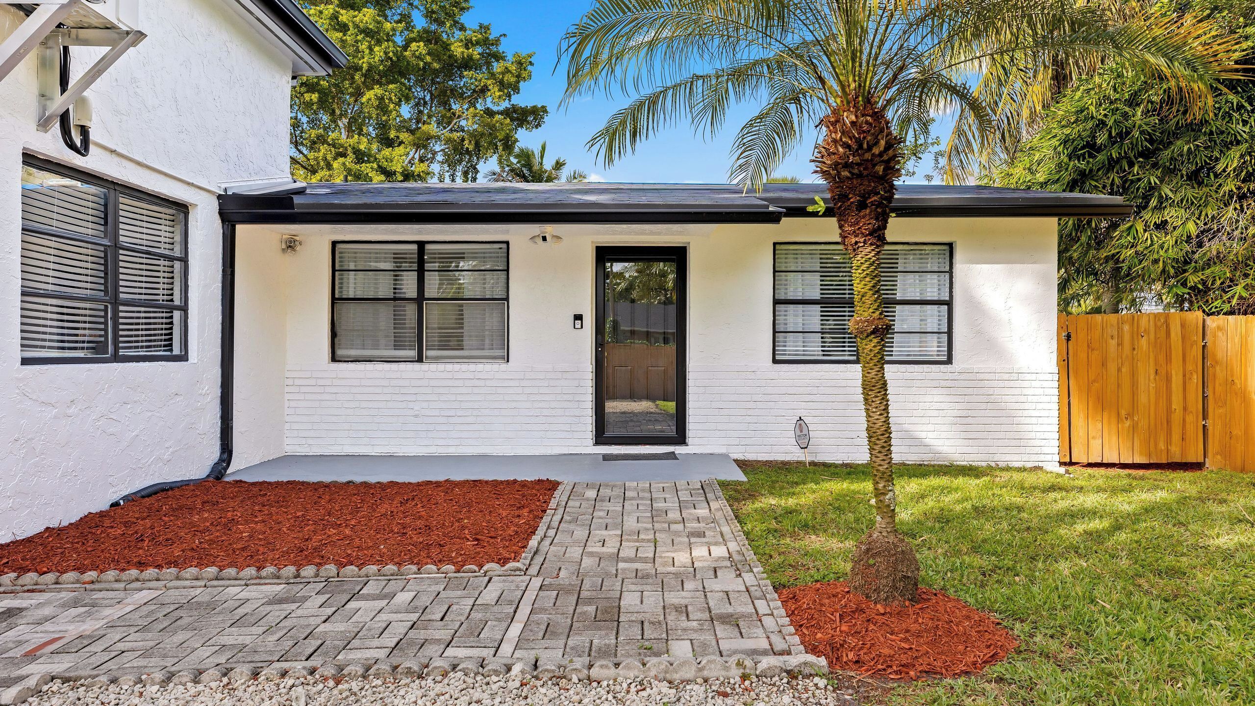 A modern two-story house with a black door, white exterior, and a small front yard with a palm tree and red mulch.
