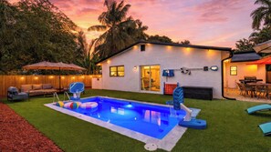 A backyard with a swimming pool, a covered patio, and a house with a door and windows. - Hoop & Splash House by Avantstay Game Room + Pool + Firepit + Minutes to Las Olas (Fort Lauderdale)
