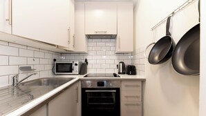 Cuina compacta amb armaris blancs, un rentapeus d'acer inoxidable, un microones, una tetera i un forn integrat.