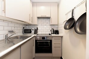 A compact kitchen with white cabinets, a stainless steel sink, a microwave, a kettle, and a built-in oven. - Bright 1BR Near Notting Hill & Portobello - Pass the Keys (London)
