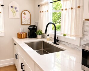 Una cucina moderna con lavello in acciaio inossidabile, mobili bianchi, una macchina da caffè e una finestra con tende.