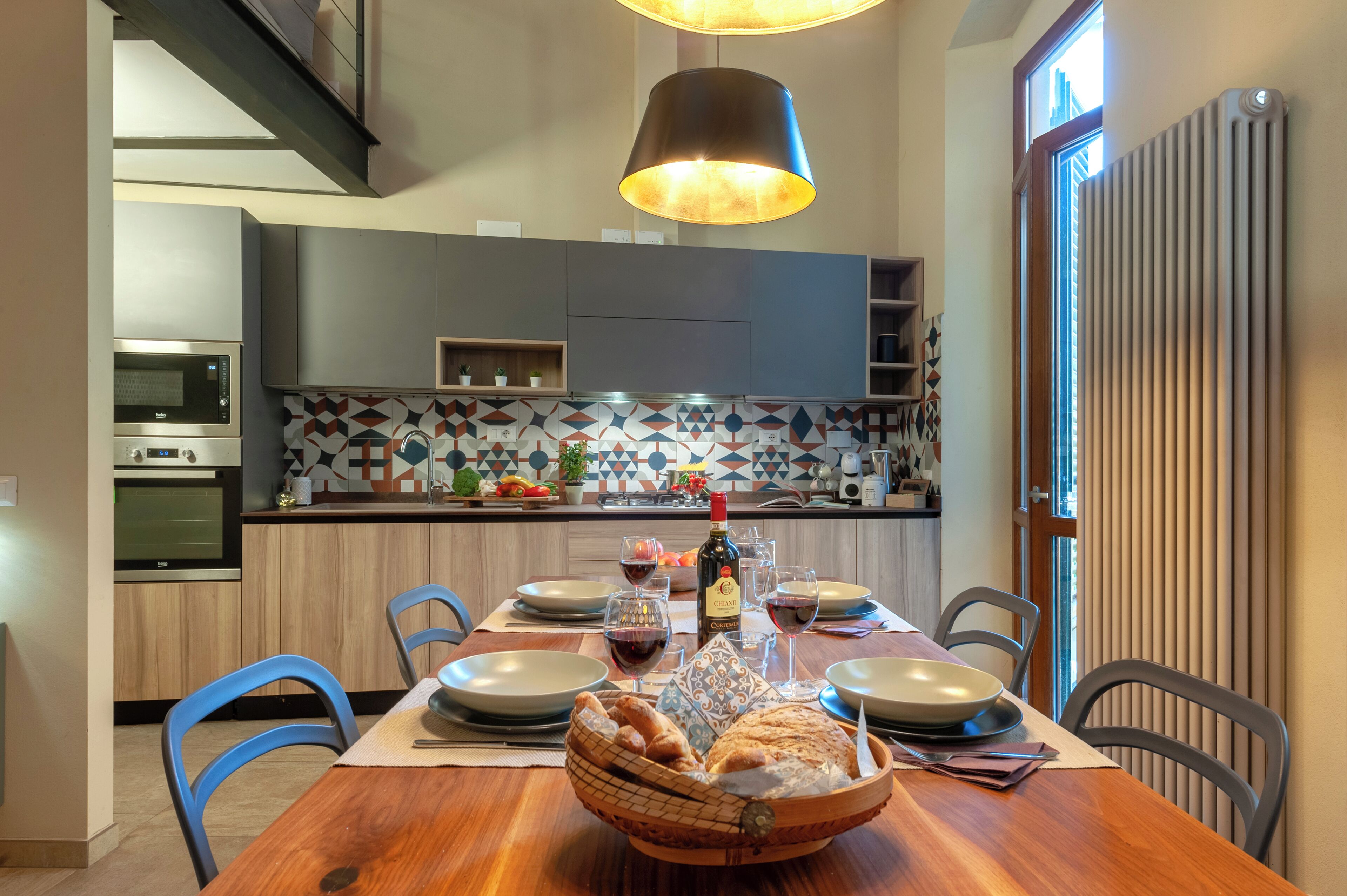 A dining area with a wooden table set for four, a basket of bread, wine glasses, and a bottle of wine.