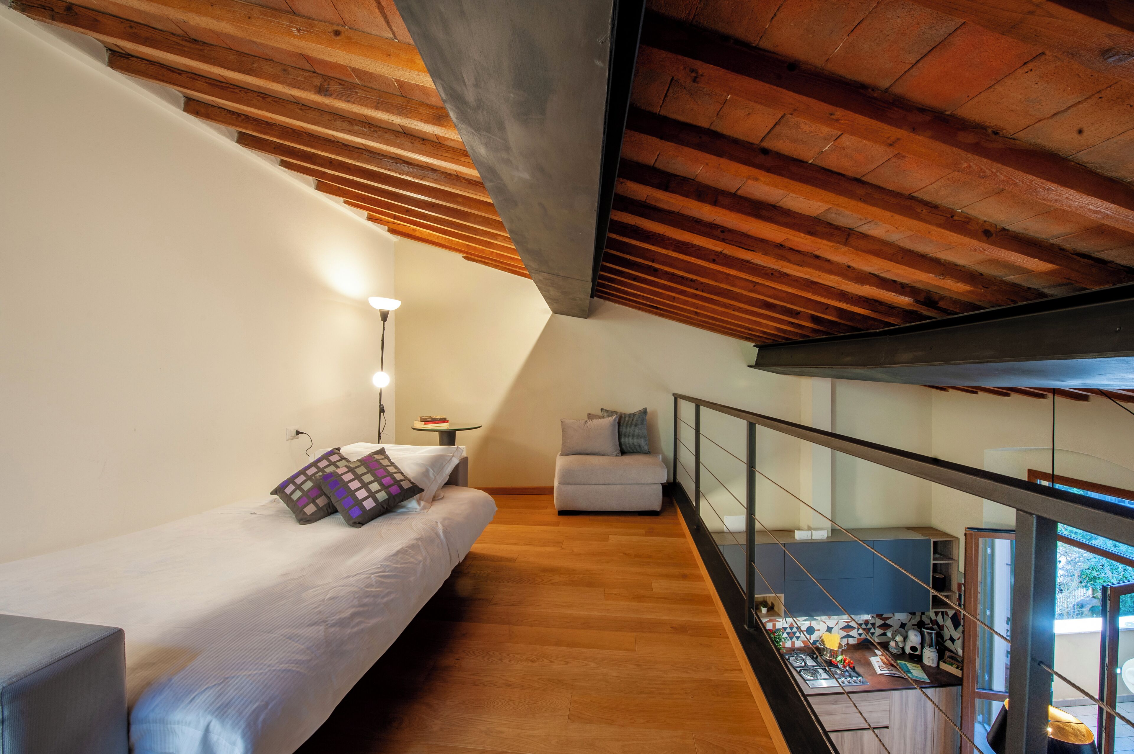 Un dormitorio moderno con techo de madera, una cama con almohadas estampadas, un pequeño sofá y vistas a una cocina y un balcón.