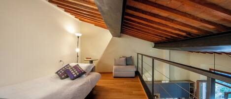 Un dormitorio moderno con techo de madera, una cama con almohadas estampadas, un pequeño sofá y vistas a una cocina y un balcón.
