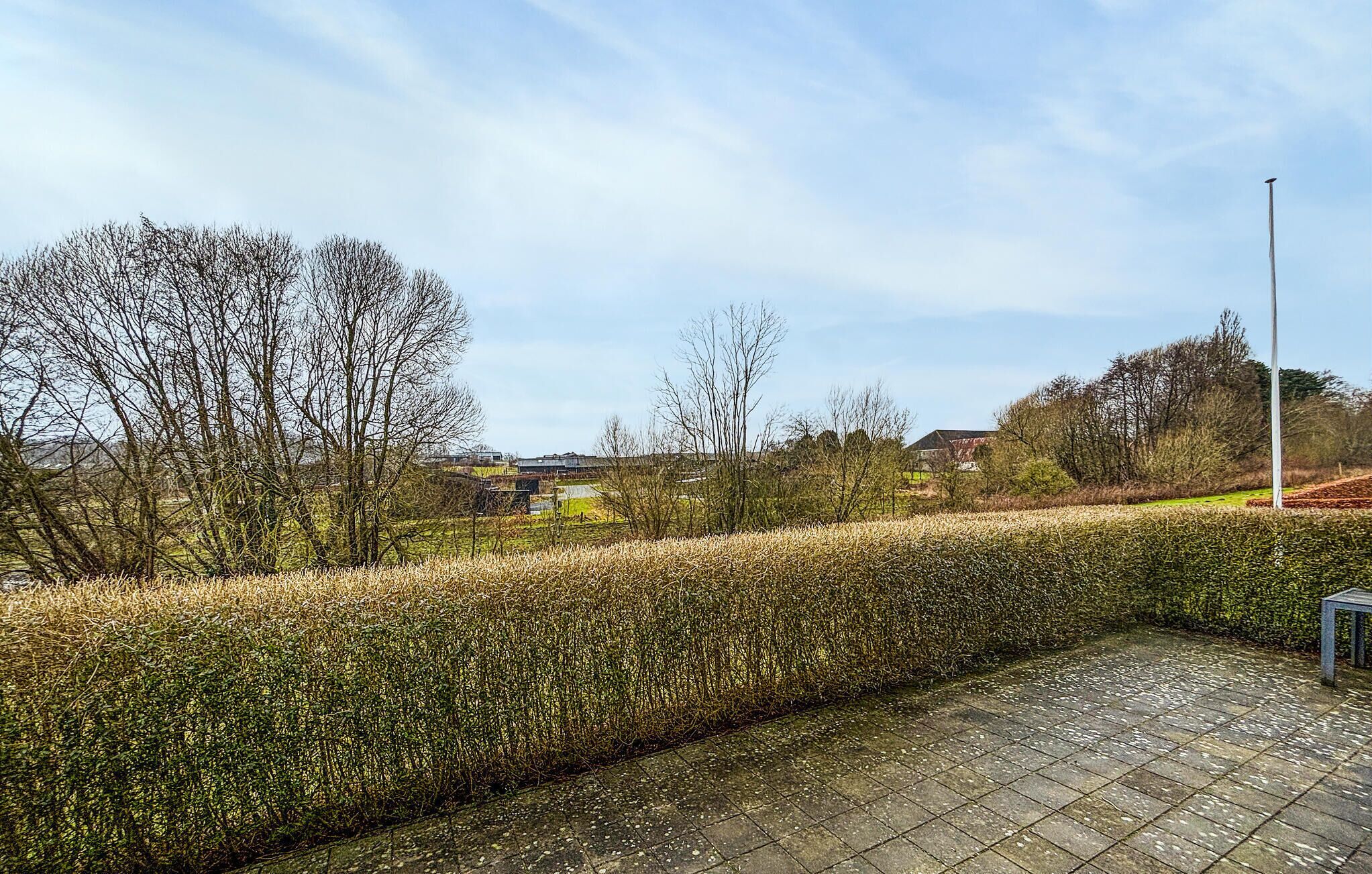 Un chemin pavé longe un arbuste taillé avec soin, et on y trouve un poteau en métal et un banc sur la droite.