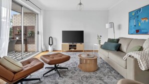 A modern living room with a sofa, a brown leather chair, a round wooden coffee table, a television, and a sliding glass door leading to an outdoor area with plants. - Coogee Oasis Ocean Outlooks near the Beach (Coogee)