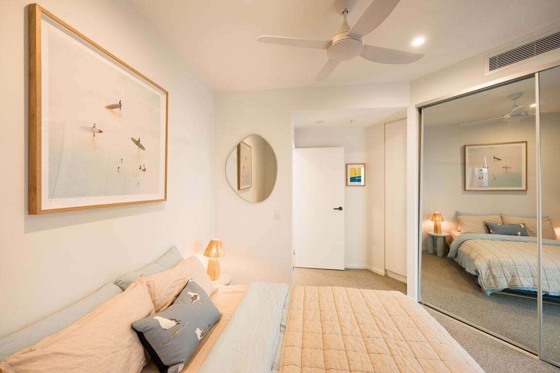Un dormitorio con una cama, una mesita de noche con lámpara, un espejo y una obra de arte enmarcada en la pared.