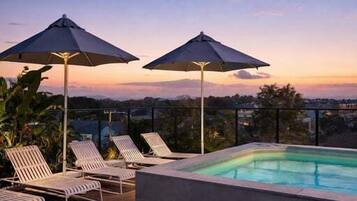 Un espace piscine aménagé sur le toit, avec des chaises longues et des parasols, entouré d’une terrasse en bois et offrant une vue sur le paysage urbain.