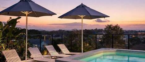 Un área de piscina en la azotea con sillones y sombrillas, rodeada por una terraza de madera y con vistas a la ciudad.