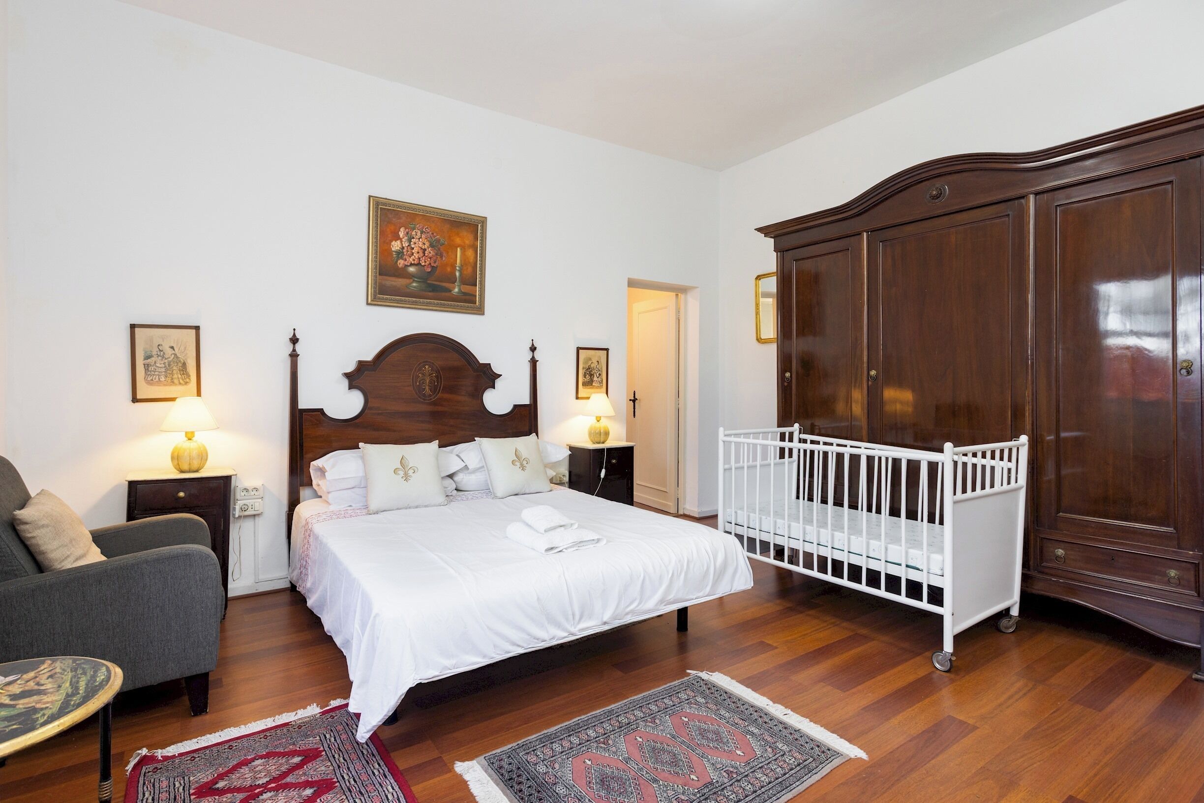 A bedroom with a wooden bed, a crib, a chair, a nightstand, and a wardrobe.