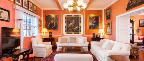 Un salon avec un téléviseur, des canapés blancs, un tapis à motifs et des portraits encadrés accrochés aux murs.