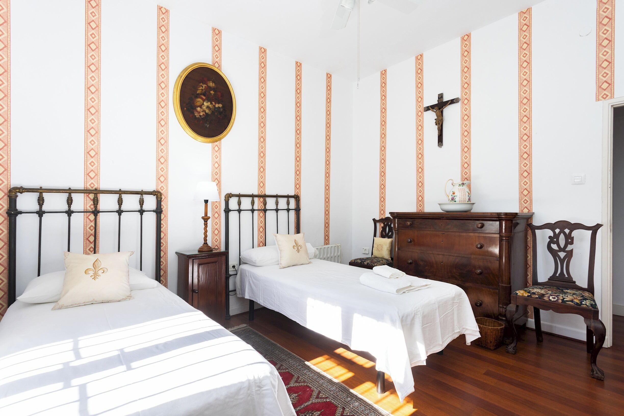 A bedroom with two beds, a wooden dresser, a chair, and a crucifix on the wall.