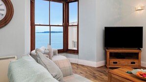A living room with a large window, a flat-screen TV, and a wooden coffee table. - 6 Paragon Court - Sea Front Apartment (Tenby)