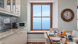 A kitchen with a view of the sea, a dining area with breakfast set, and a clock on the wall. - 6 Paragon Court - Sea Front Apartment (Tenby)