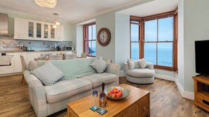 A living room with a sofa, armchairs, a coffee table with a fruit bowl, and a TV stand. - 6 Paragon Court - Sea Front Apartment (Tenby)