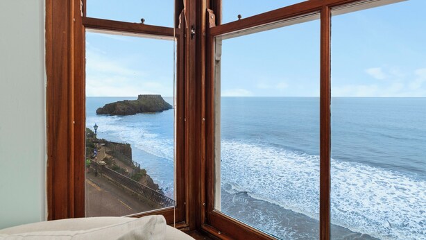 A room with a large window overlooking a rocky coastline and the sea. - 6 Paragon Court - Sea Front Apartment (Tenby)