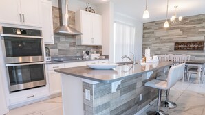 A modern kitchen with stainless steel appliances, a large island with a bowl sink, and a dining area with a wooden wall and hanging lights. - Costa Championsgate 4BDR - Quiet House with Jacuzzi (Davenport)