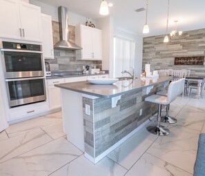 A modern kitchen with stainless steel appliances, a large island with a bowl sink, and a dining area with a wooden wall and hanging lights. - Costa Championsgate 4BDR - Quiet House with Jacuzzi (Davenport)