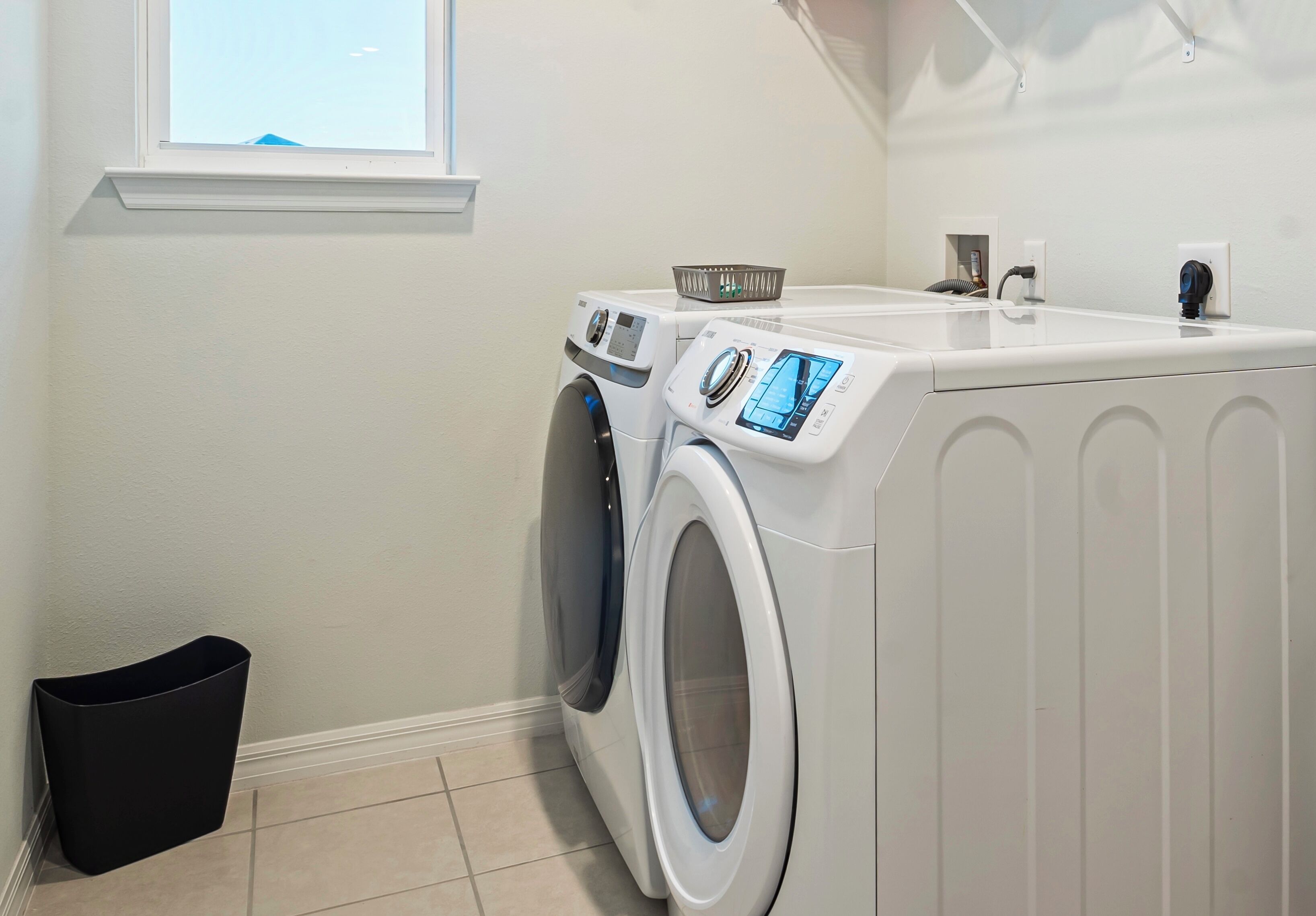 A laundry area with two front-loading washing machines, a drying rack, and a black trash bin.
