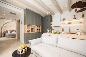 A modern living room with a white sofa, a wooden coffee table with fruit, a kitchen area with a countertop and shelves, and a stone wall in the background. - Villa Analidia Comfort (Koridallos)