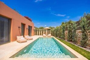 Une piscine avec des chaises longues, un espace de détente couvert et un mur de pierre agrémenté d’arbres.