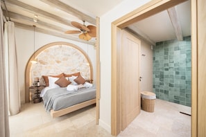 A bedroom with a stone wall, a bed with pillows, a wooden door, and a bathroom with a tiled wall. - Villa Analidia Comfort (Koridallos)