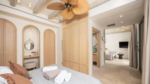 Ein Schlafzimmer mit einem Bett, einem Deckenventilator, einem Holzschrank, einem Spiegel und einem durch eine offene Tür einsehbaren Badezimmer.